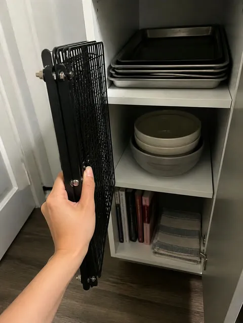 Folded three-tier cooling rack being stored beside bakeware in a small cabinet