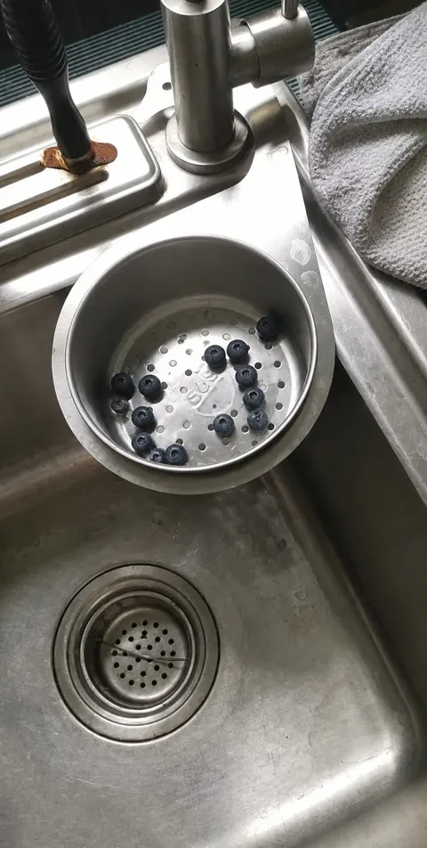 Casual phone photo of the stainless sink strainer holding blueberries over a kitchen sink