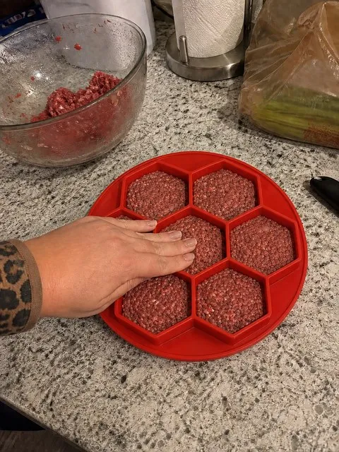 Round red seven-cavity silicone patty mold with raised hexagonal walls being filled on a home kitchen counter