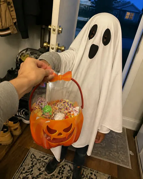 Child in a ghost costume receiving an orange pumpkin treat bag