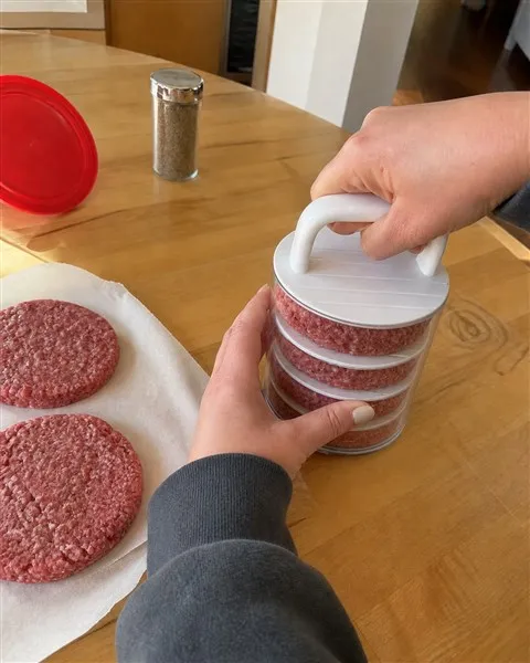 Home cook pressing and stacking evenly shaped burger patties in a closed clear cylinder on a wood kitchen table