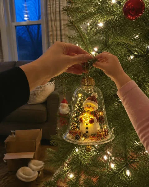 Parent and child hanging a snowman light-up bell ornament on a tree