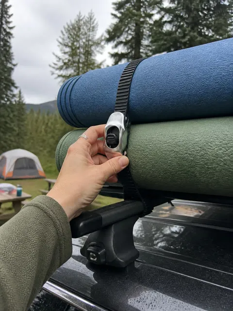 Hand checking a utility strap around camping mats on a roof rack