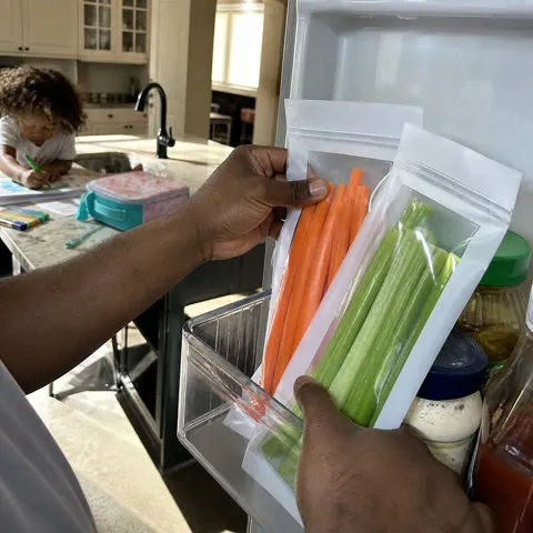 Carrots and celery in long clear bags being placed in a family refrigerator